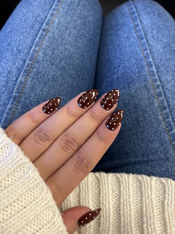 Close-up of a hand showing manicured nails painted in a brown color with white polka dots, resting on a knitted sweater and denim fabric.