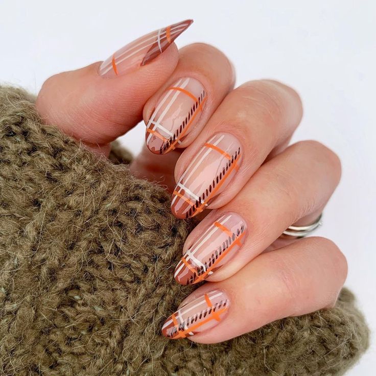Close-up of a hand showing stylish long nails with a nude base color and geometric patterns in orange and black, resting on a knitted brown sweater.