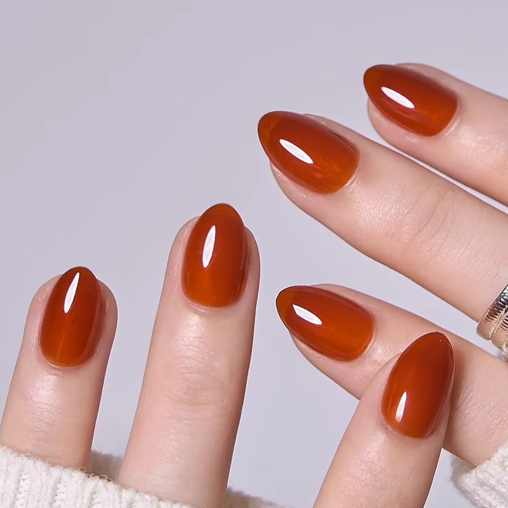 Close-up of hands showcasing neatly manicured fingernails painted in a glossy burnt orange color, featuring various nail shapes.