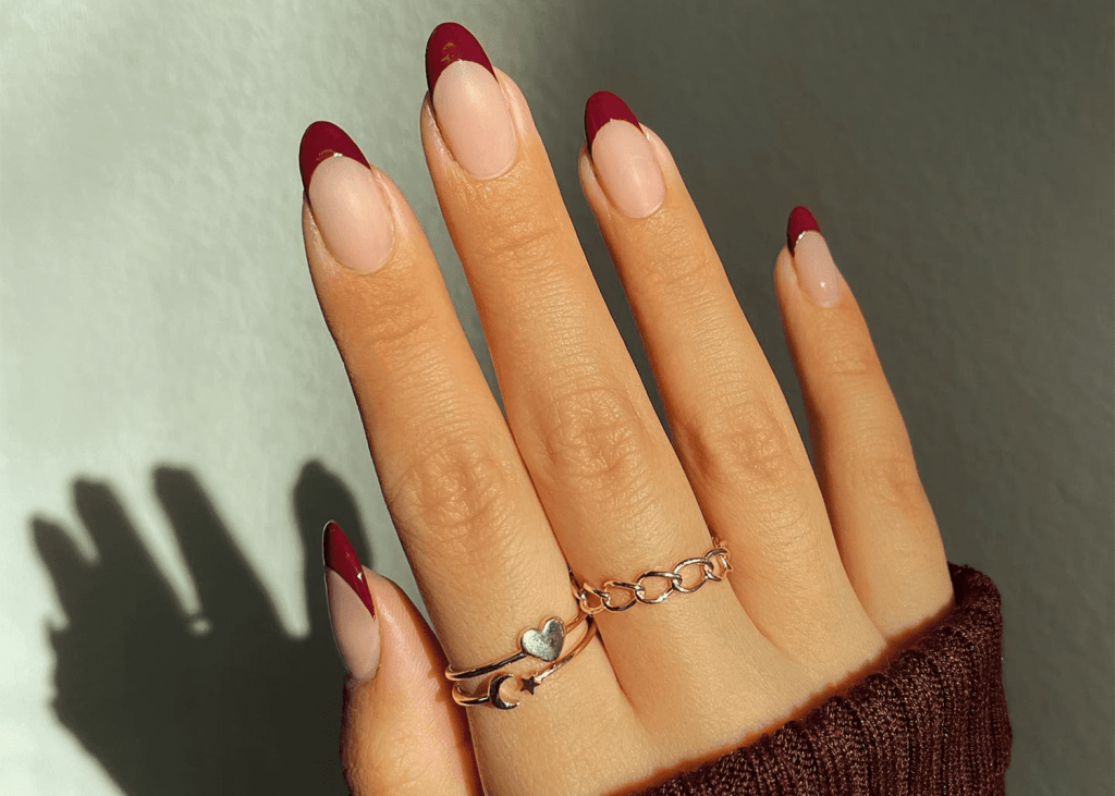Close-up of a hand with long, almond-shaped nails featuring a burgundy French tip design. The hand is adorned with multiple rings, including a heart-shaped and a chain-style ring. The background is softly lit, highlighting the elegant manicure.