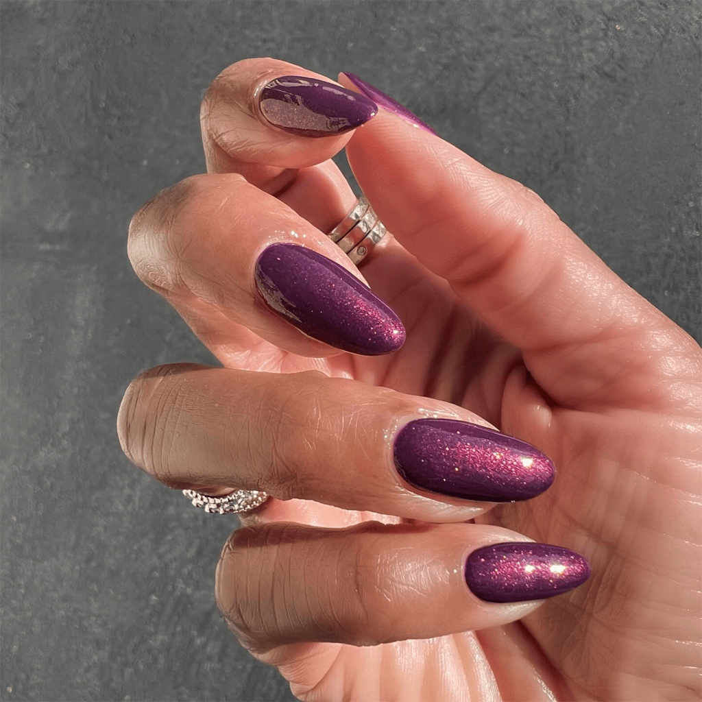 Close-up of a hand displaying long, purple glitter nail polish on the fingers, with rings on the fingers and a textured background.