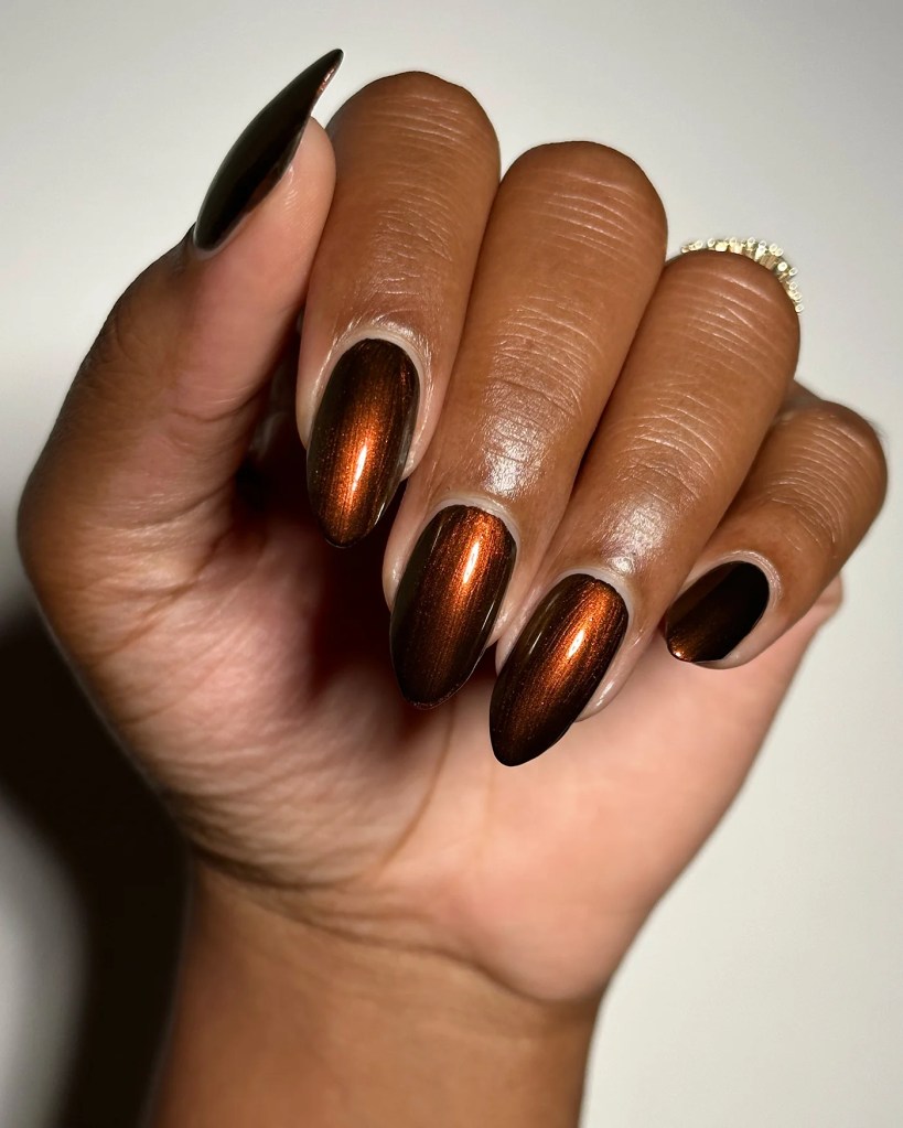 A close-up of a hand showcasing long, almond-shaped nails with a gradient from dark to shiny copper brown. The nails have a glossy finish, and the hand is positioned against a light background.