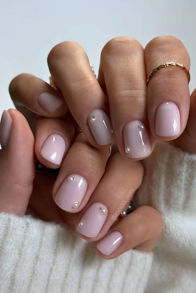 Close-up of well-manicured hands with pale pink nails featuring small decorative gems, wearing minimalist rings, and resting on a soft, textured fabric.