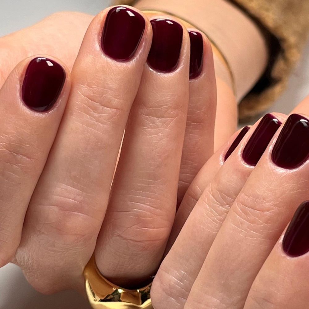 Close-up of hands with neatly manicured nails painted in a deep burgundy color, featuring a glossy finish. One hand is visible wearing a gold ring.
