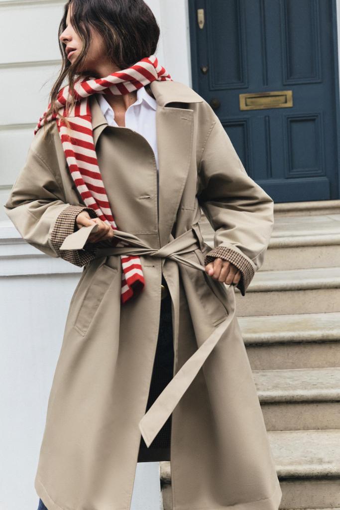 A woman wearing a beige trench coat and a red and white striped scarf stands on stairs, showing a stylish pose.