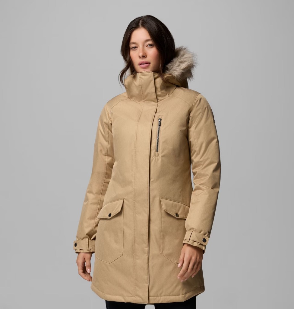 A model wearing a beige winter coat with a fur-lined hood, standing against a gray background.