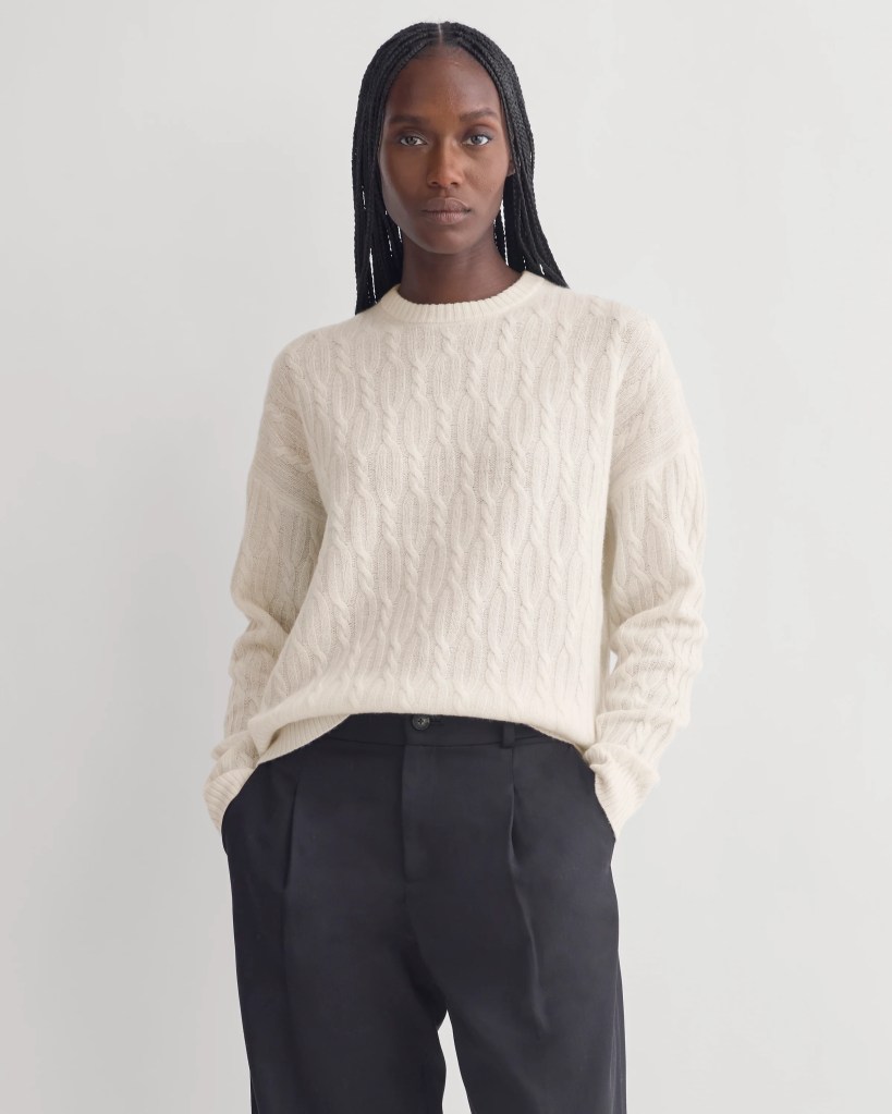 A model wearing a cream-colored cable knit sweater paired with black pants, standing against a light gray background.