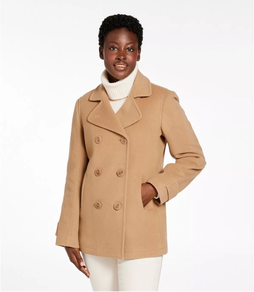 A woman wearing a stylish beige double-breasted coat with a white turtleneck and light-colored pants, posing with a confident smile against a simple white background.