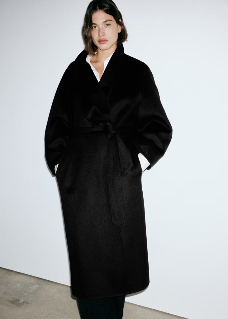 A woman wearing a black overcoat with a tie waist, standing against a minimalistic white background.
