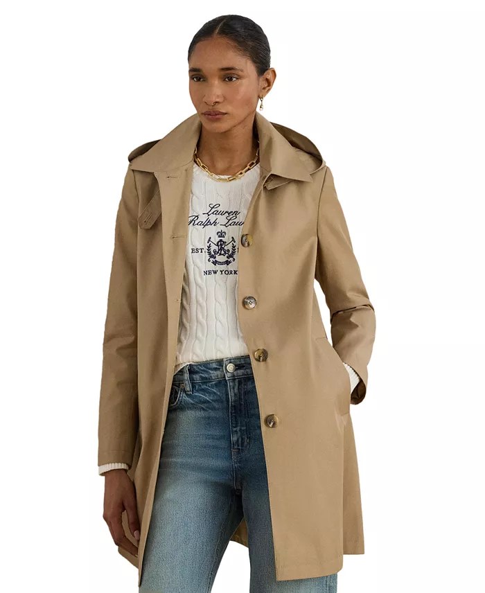 A model wearing a beige trench coat over a white knitted sweater with a logo, paired with blue jeans.