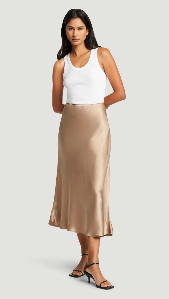 A fashionable woman wearing a white tank top and a beige satin midi skirt, standing against a neutral background.
