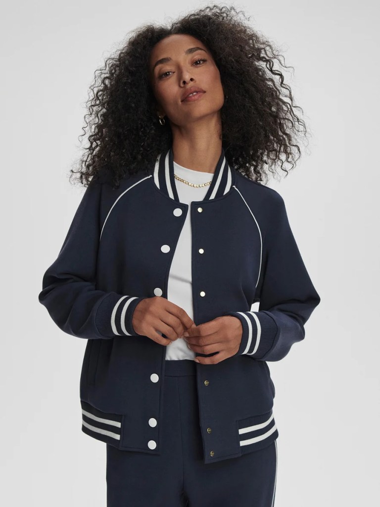 A woman with curly hair wearing a navy blue varsity jacket with white stripes on the cuffs, standing confidently with her hands together.