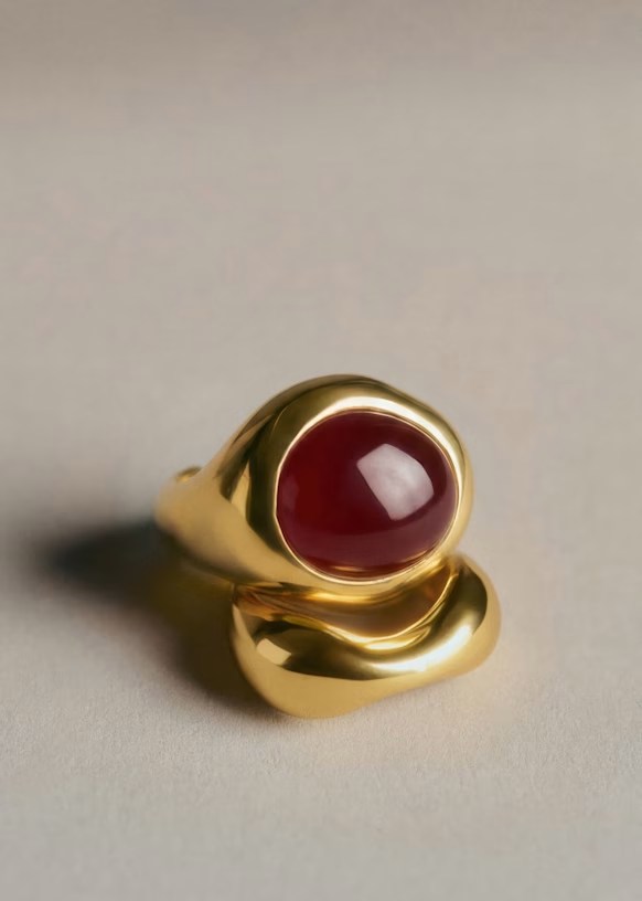 A gold ring featuring a smooth, round red stone set on top, displayed against a neutral background.