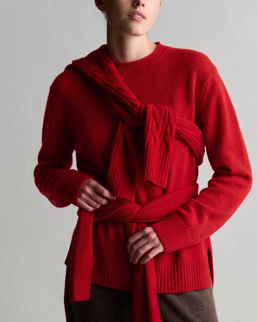 A woman wearing a red sweater with a stylish twist feature, showcasing a knitted design wrapped around her torso.