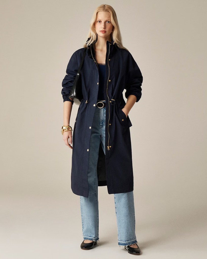 A woman wearing a navy blue long jacket, paired with a black top and light blue jeans, standing confidently against a neutral background.