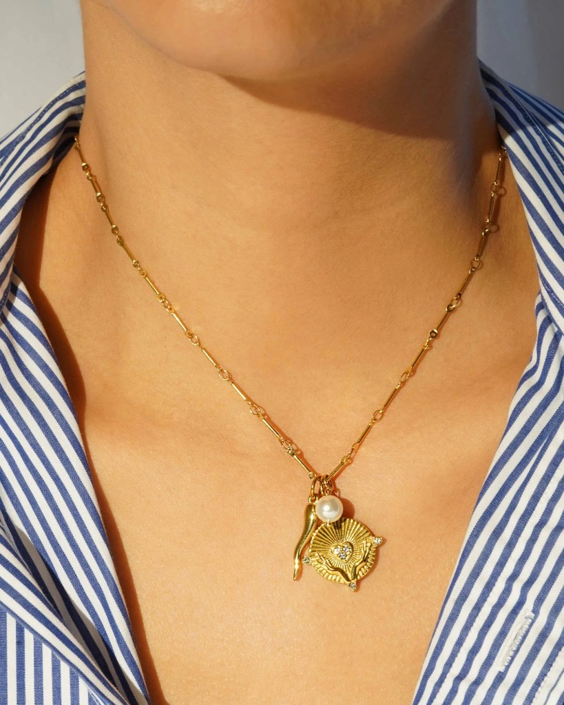 Close-up of a person wearing a gold necklace with a floral pendant and a pearl, against a background of a blue and white striped shirt.