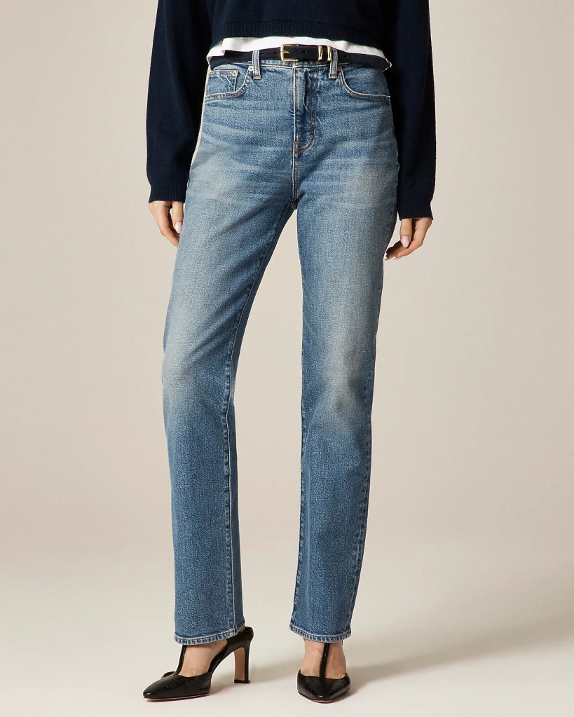 A model wearing high-waisted blue jeans paired with a dark sweater and black high-heeled shoes, standing against a neutral background.