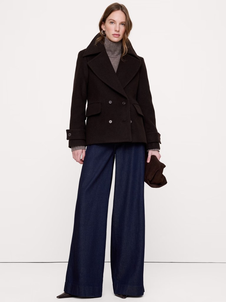 A woman wearing a dark peacoat and a gray turtleneck, paired with wide-leg denim jeans, stands confidently with one hand on her hip and holding a brown hat in the other.