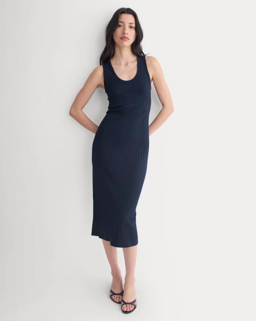A woman wearing a sleeveless navy ribbed midi dress, standing with one hand on her hip and the other relaxed at her side, against a light gray background.