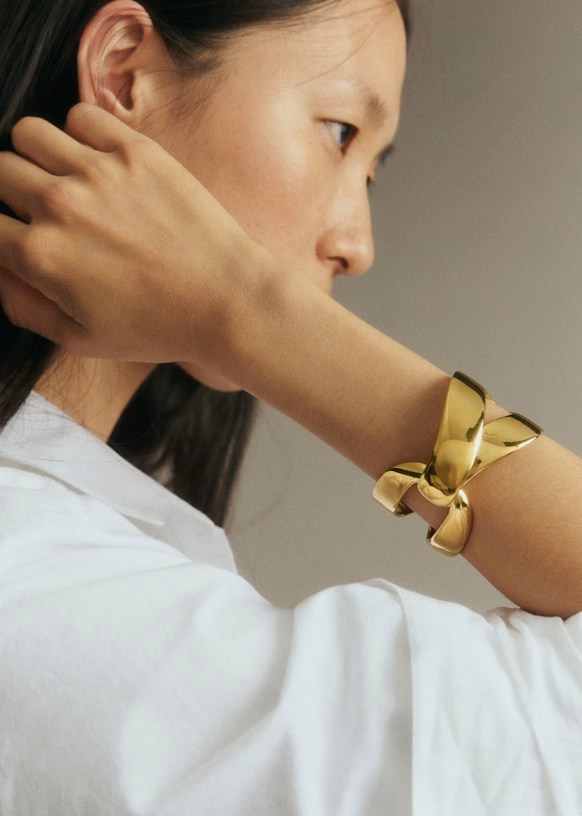 A close-up of a woman's wrist adorned with a shiny gold bracelet featuring a knot design, with her hand resting gently on her collarbone.