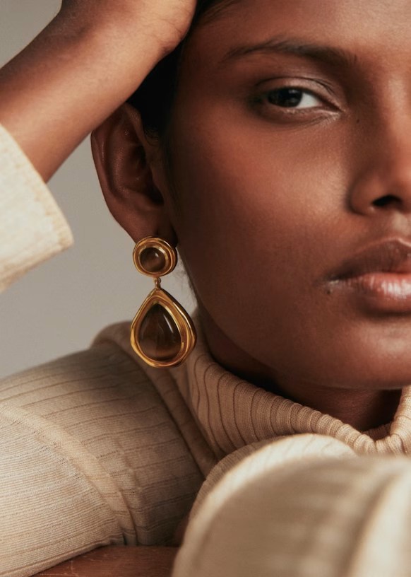 Close-up of a woman wearing elegant gold earrings, featuring a teardrop design, against a muted background. Her hand is gently resting on her head, showcasing her textured skin and neutral-toned outfit.