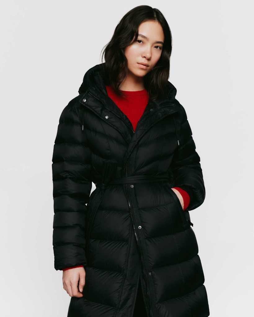 A young woman wearing a black puffer coat with a hood, standing against a plain white background. She has long, wavy hair and is wearing a red sweater underneath the coat.