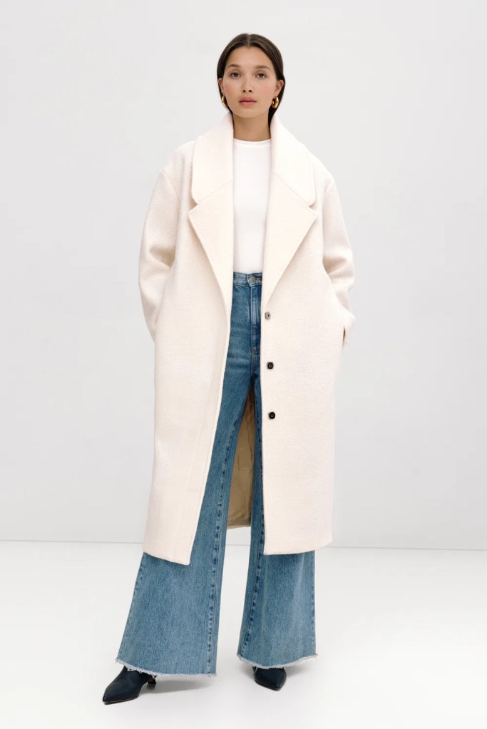 A woman stands confidently in a light-colored oversized coat over a white top and blue wide-leg jeans, paired with black ankle boots, set against a neutral background.