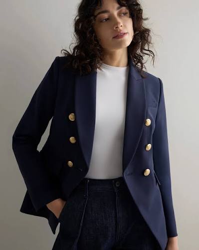 A woman wearing a navy double-breasted blazer with gold buttons, paired with a white shirt and dark jeans, standing against a light background.