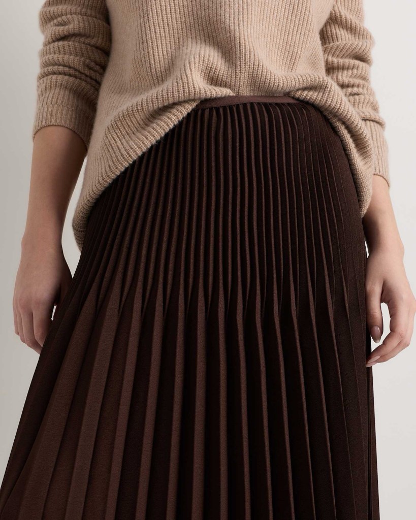 A close-up of a woman wearing a beige ribbed sweater and a brown pleated skirt.