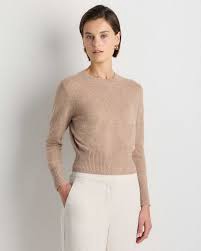 A woman wearing a light beige long-sleeve sweater paired with light-colored trousers, standing against a plain background.