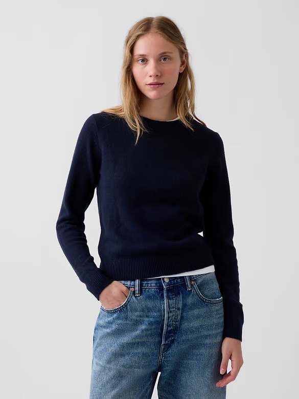A young woman standing in front of a plain light background, wearing a dark navy sweater and faded blue jeans, with her hair down and a neutral expression.