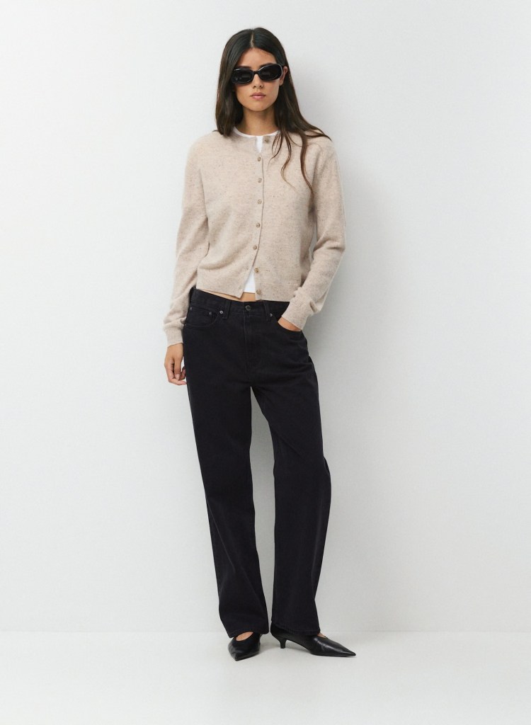 A woman wearing a beige cardigan over a white shirt, paired with black wide-leg pants and black pointed-toe shoes, stands against a plain background. She has long dark hair and is wearing sunglasses.