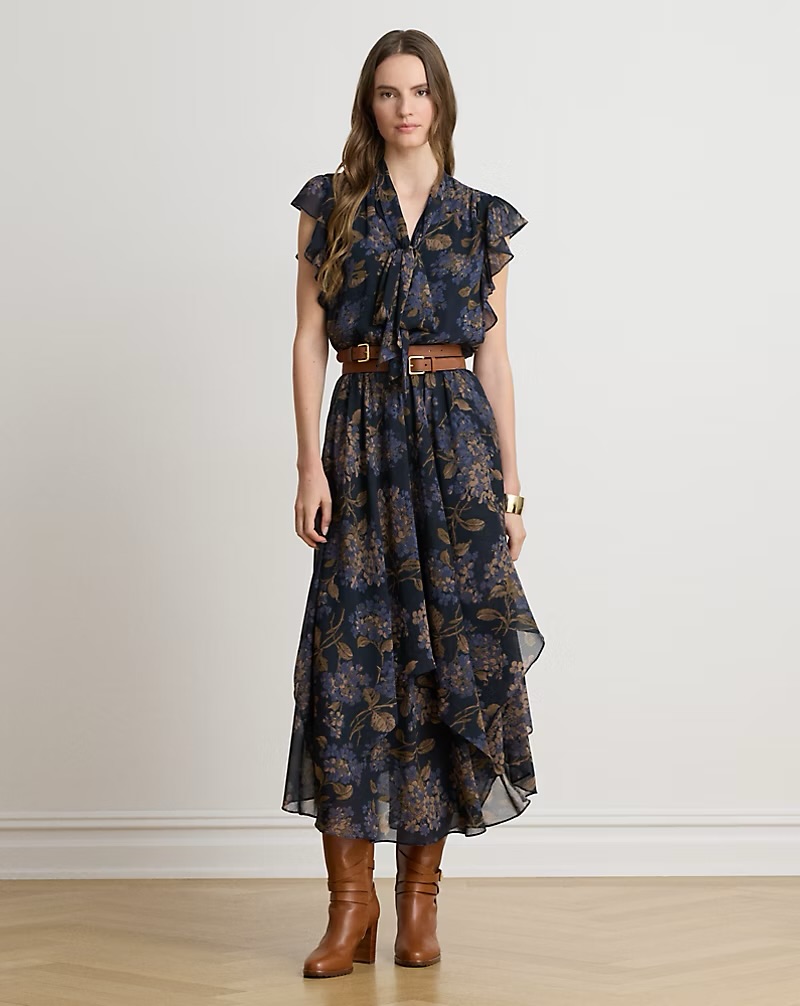 A woman wearing a floral maxi dress with a ruffled neckline, belted at the waist, standing in brown leather boots.