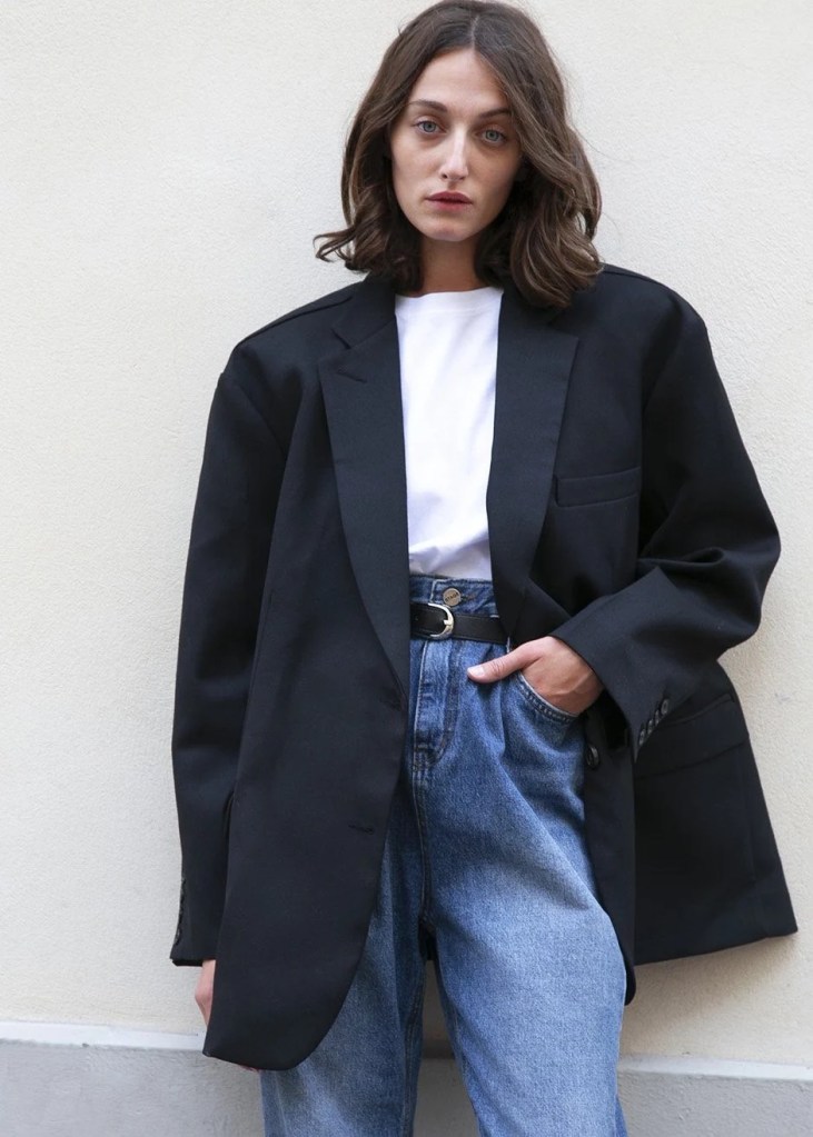 A model wearing an oversized black blazer over a white t-shirt, paired with high-waisted blue jeans, posing against a neutral-colored wall.