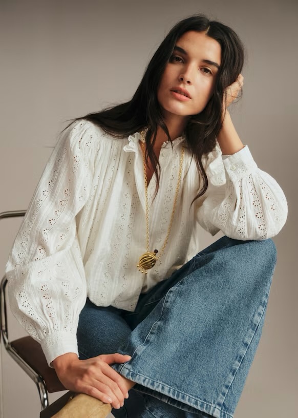 A woman with long dark hair wearing a white blouse with intricate detailing and blue flared jeans, sitting in a relaxed pose with a gold necklace.