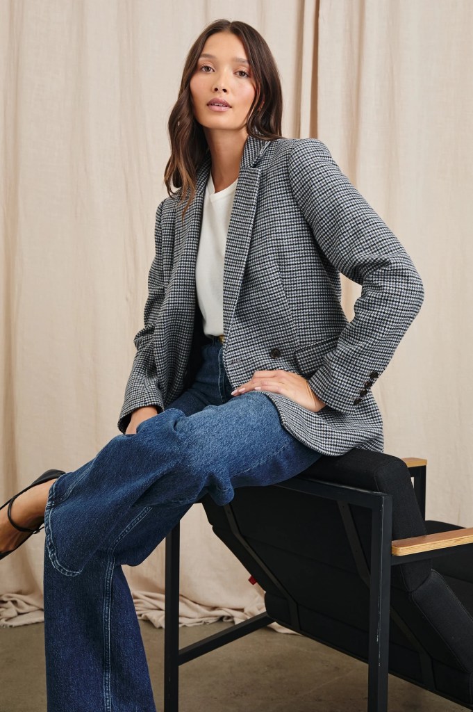 A woman sitting on a chair wearing a stylish plaid blazer over a white top and denim jeans, set against a beige backdrop.