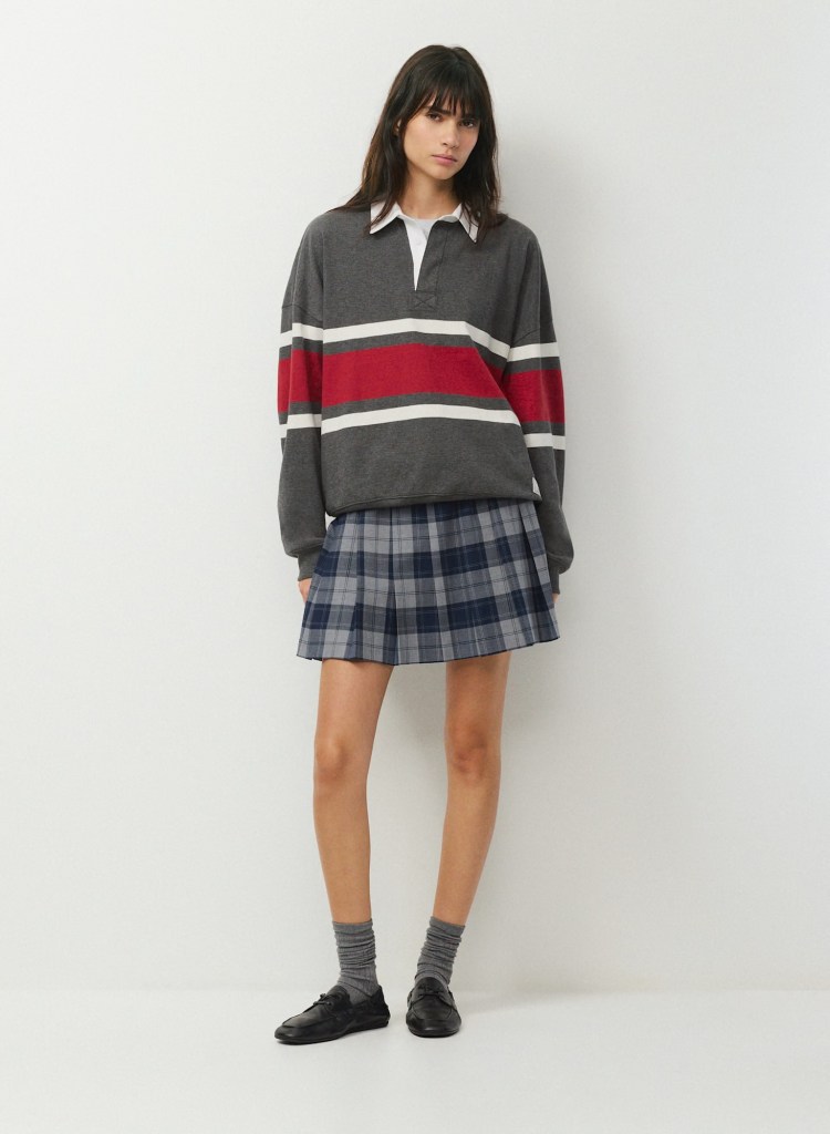 A young woman standing against a plain background, wearing a gray long-sleeve shirt with red and white stripes, a white collared shirt underneath, and a plaid skirt. She is also wearing gray socks and black shoes.