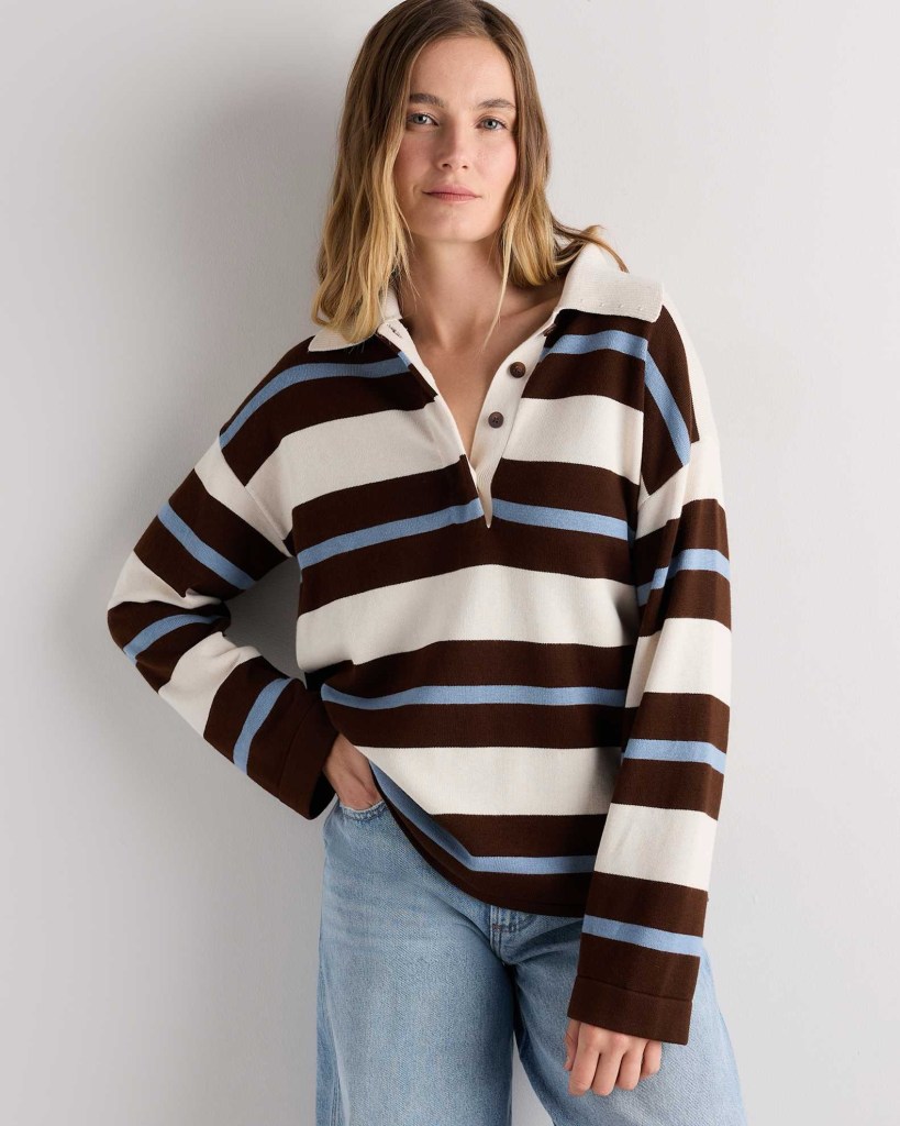 A woman standing against a light wall, wearing a striped long-sleeve shirt in brown, white, and light blue, paired with light blue jeans.