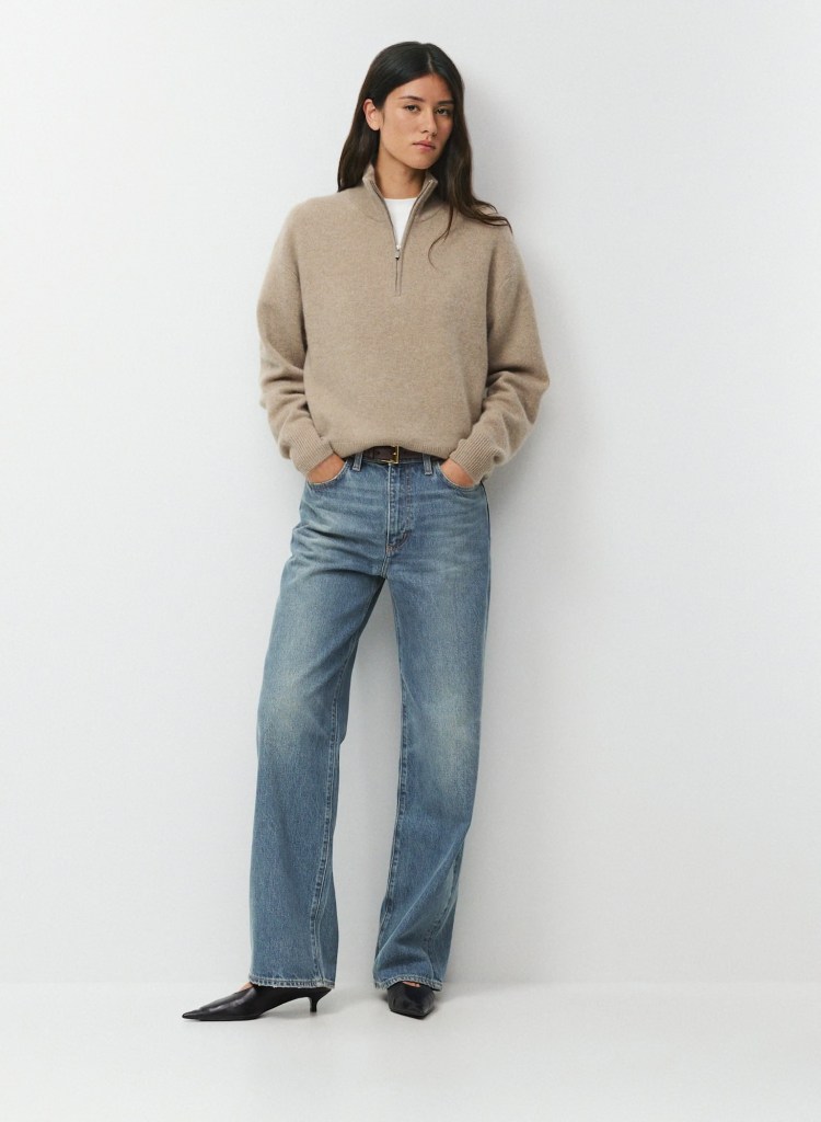 A model wearing a beige half-zip sweater, white t-shirt, and loose-fitting blue jeans, standing against a plain white background.