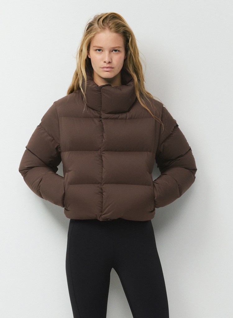 A model wearing a brown puffer jacket with a high collar, paired with black leggings, standing against a plain background.