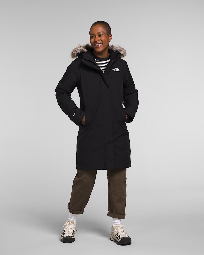 Person smiling while wearing a black winter coat with a fur-lined hood, striped shirt, and casual pants and shoes.