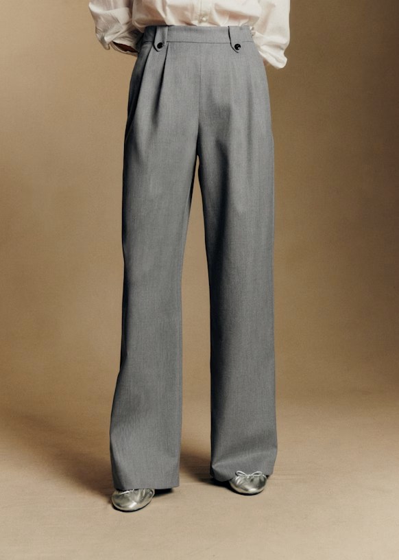 A person standing in tailored gray trousers with a high waist and pleats, paired with a light-colored, long-sleeved shirt and metallic shoes, against a neutral background.