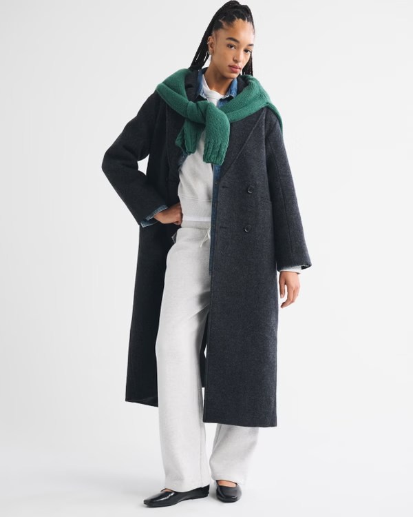 A model wearing a gray long coat over a light gray outfit, with a green scarf draped around the neck, standing against a white background.