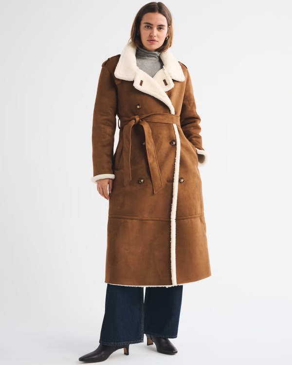 A woman wearing a long brown coat with a cream-colored collar and cuffs, styled with a gray turtleneck and wide-leg denim pants.