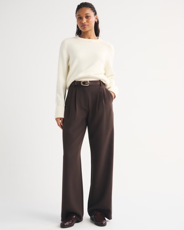 A woman standing confidently, wearing an off-white sweater and brown high-waisted trousers with a belt, posing against a plain background.