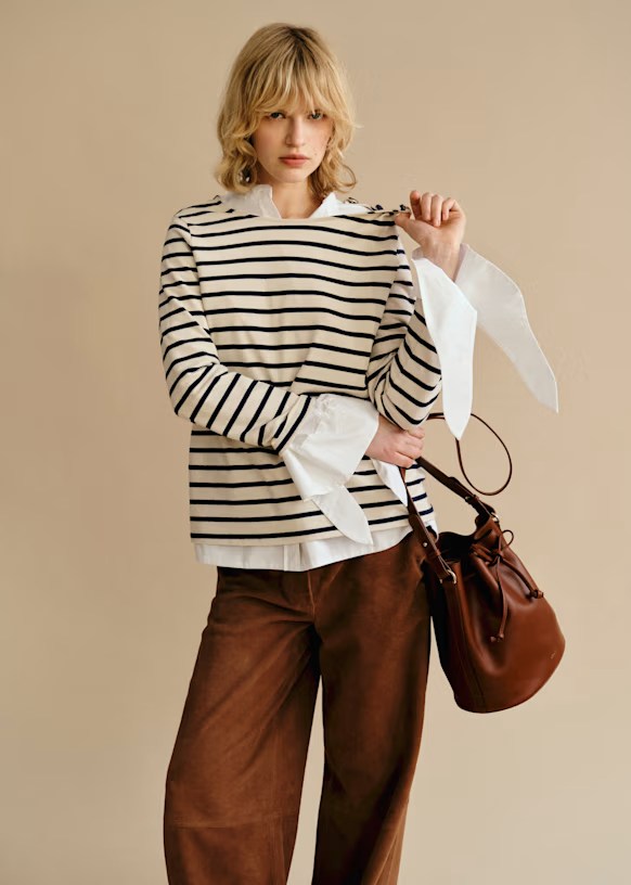 A model wearing a striped long-sleeve top with white cuffs, paired with brown wide-leg pants, holding a brown handbag against a neutral background.