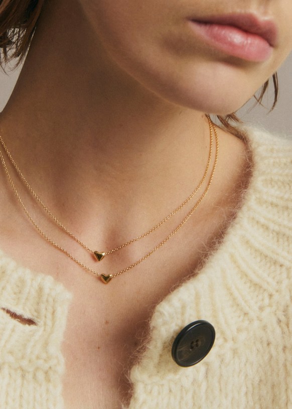 Close-up of a person's neck wearing two delicate gold necklaces with heart-shaped pendants, paired with a cozy cream-colored cardigan.