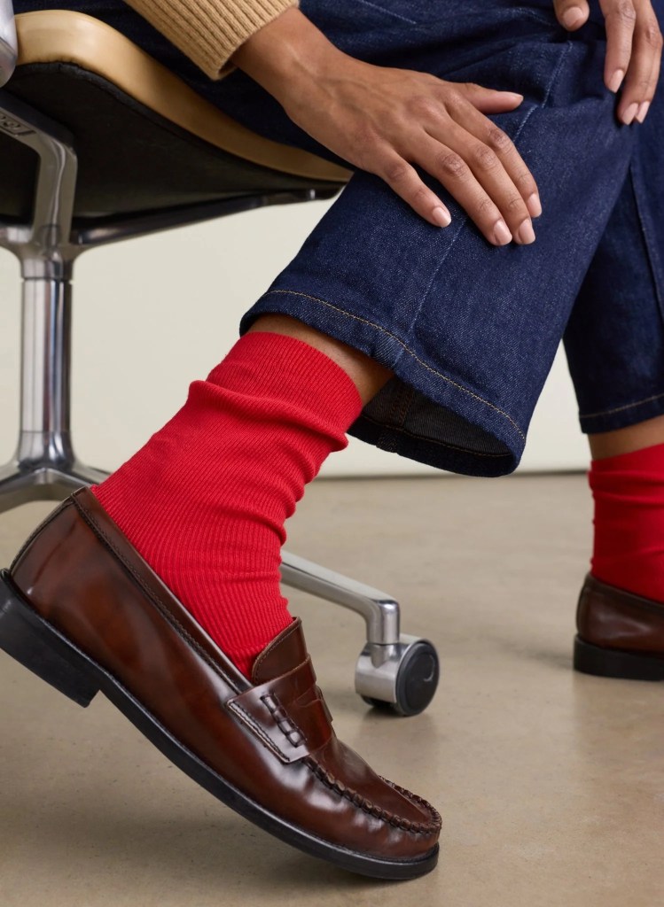 Close-up of a foot wearing a brown loafer shoe and bright red textured socks, with a hand resting on the leg, seated on a chair.