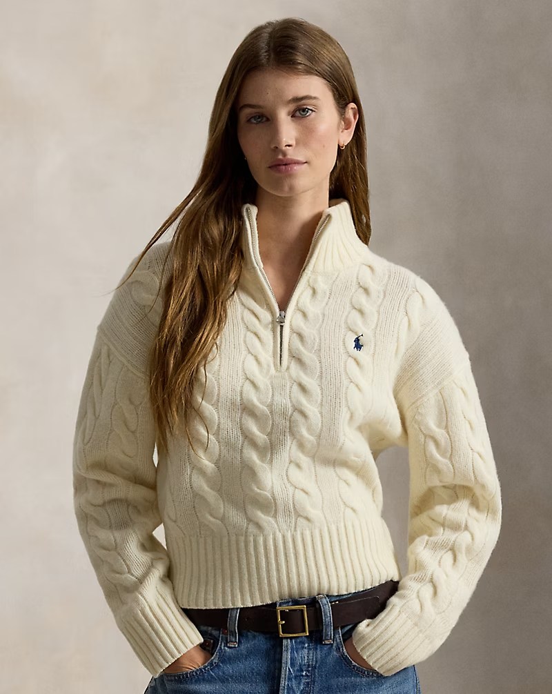 A model wearing a cream-colored cable-knit sweater with a half-zip collar, paired with high-waisted denim jeans and a brown belt.