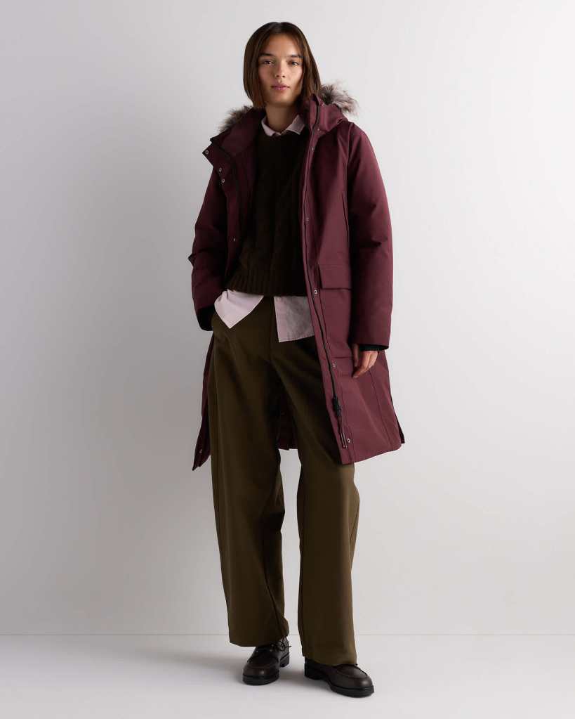 A young woman stands confidently in a maroon parka with a fur-lined hood, paired with a dark sweater, white shirt, and olive green wide-leg pants. She is wearing black shoes and posing against a neutral background.